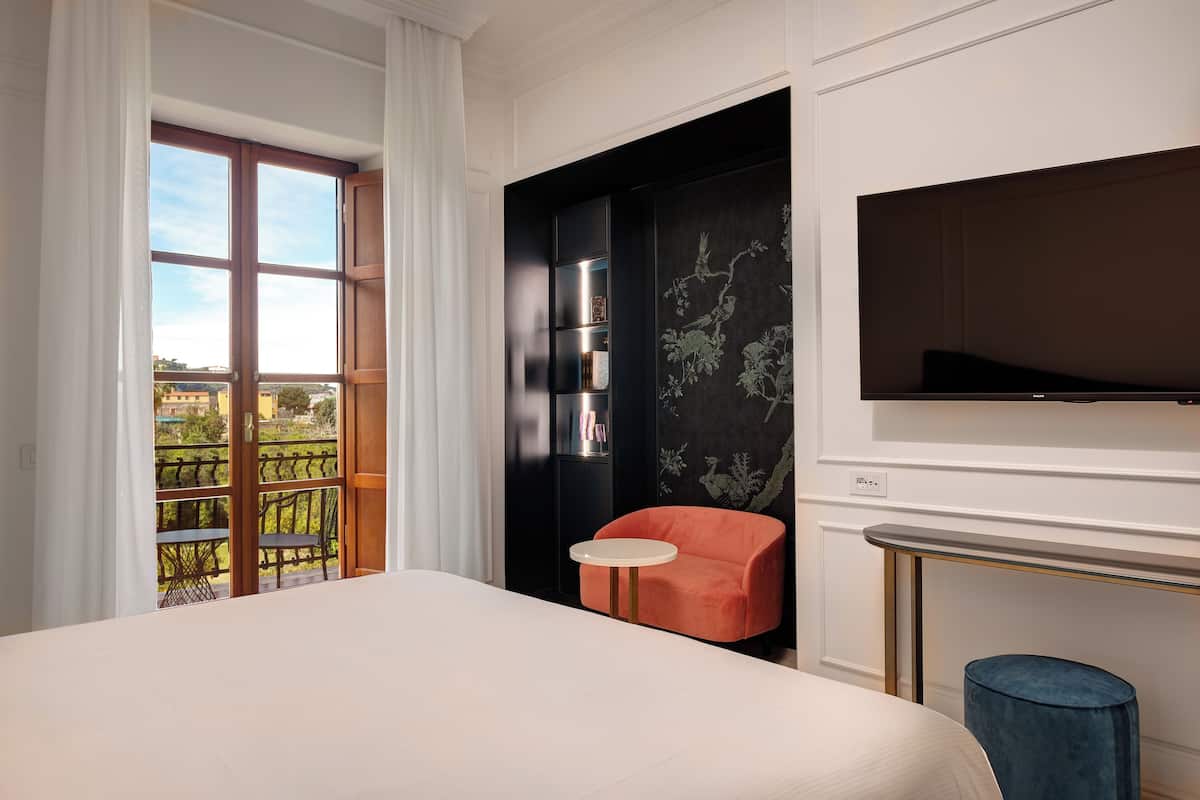 A hotel room with a large bed, a small round table, a red chair, a television, and a balcony with a railing.