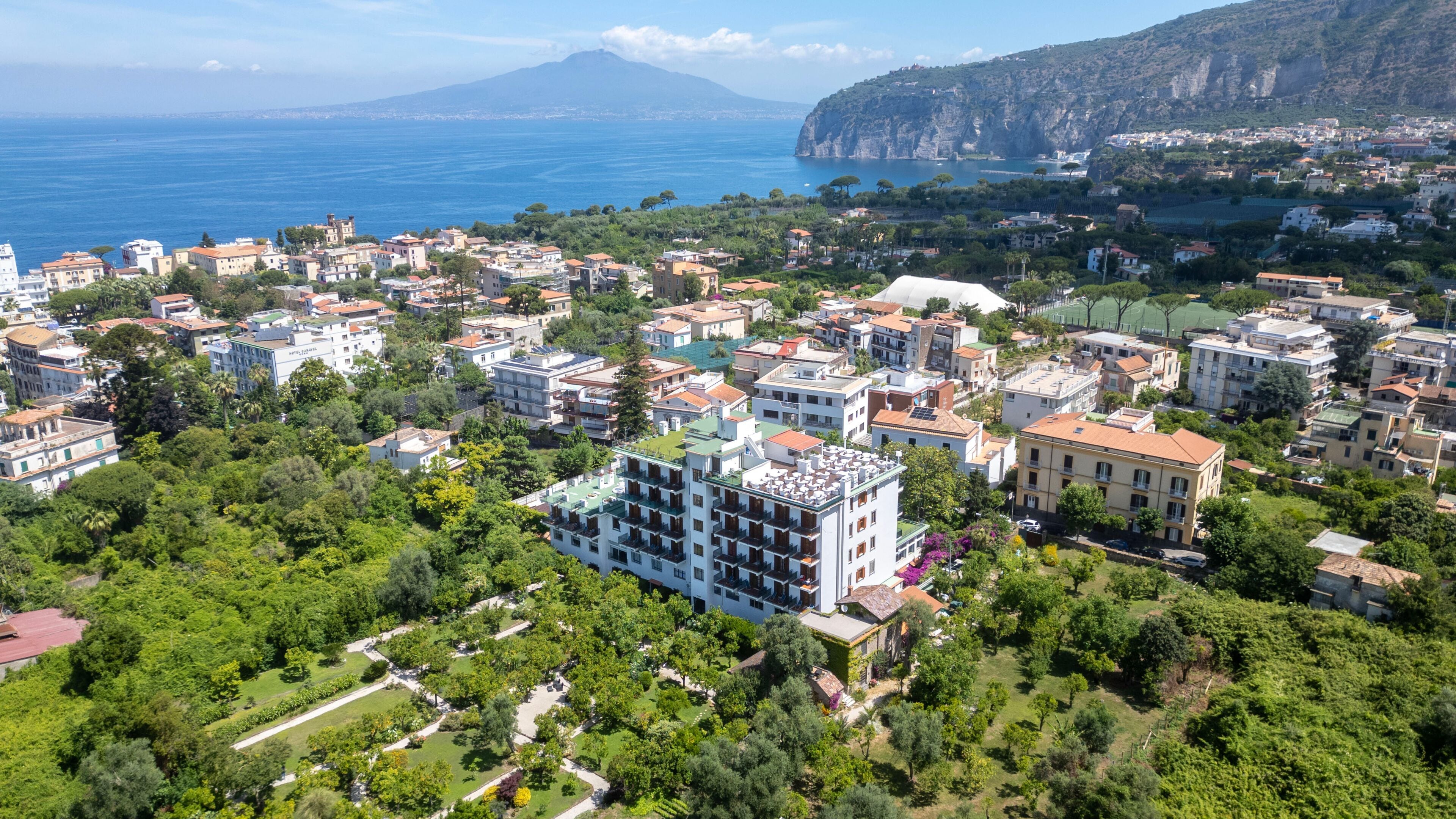A residential area with multiple buildings, green spaces, and a coastline in the background.