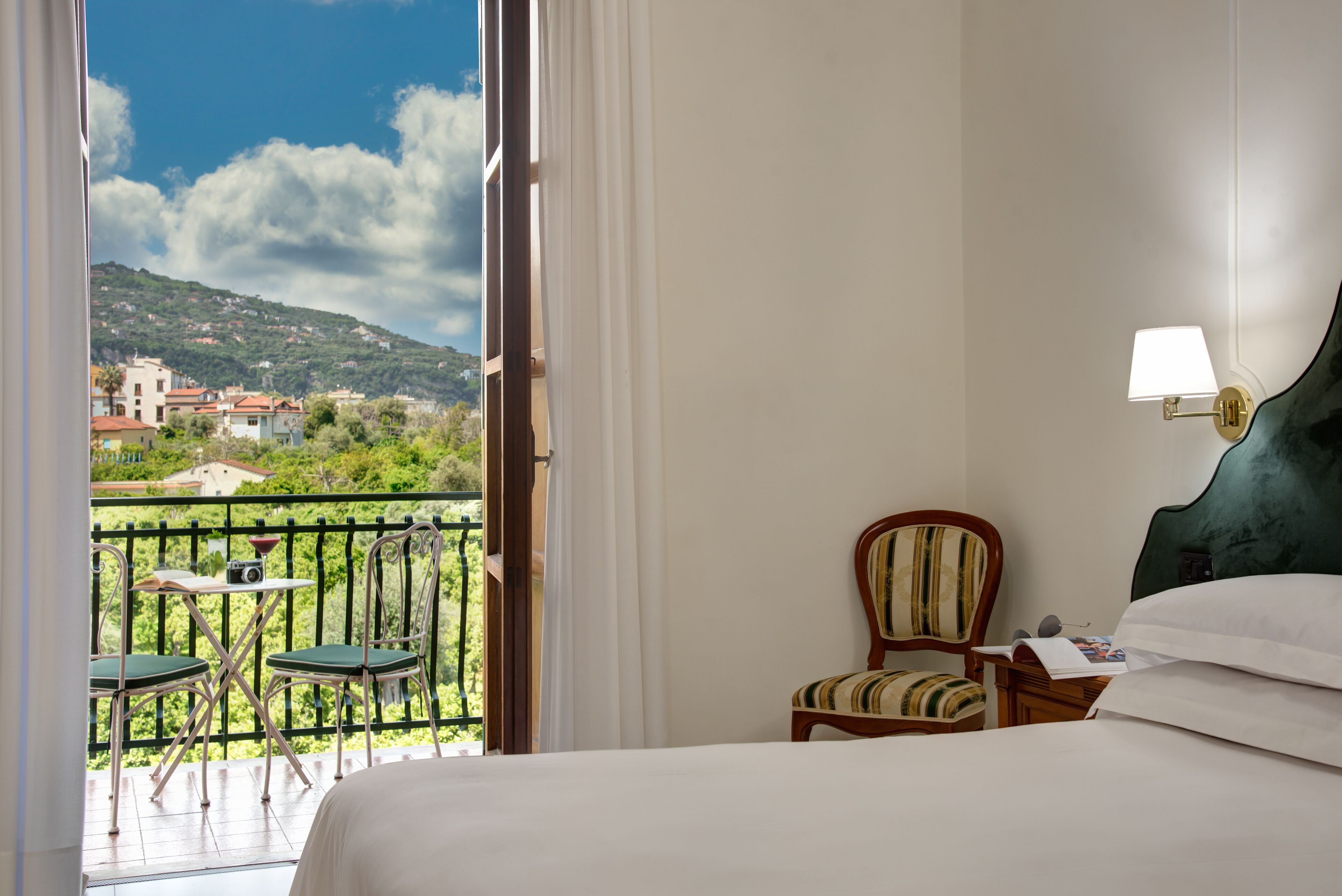 A hotel room with a balcony, a bed, a chair, and a table with a camera.