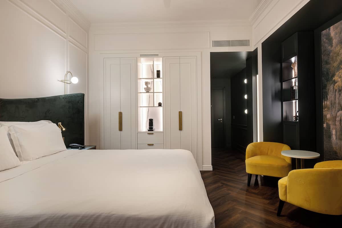 A hotel room with a bed, a wardrobe, a vanity with a mirror, and two yellow armchairs.