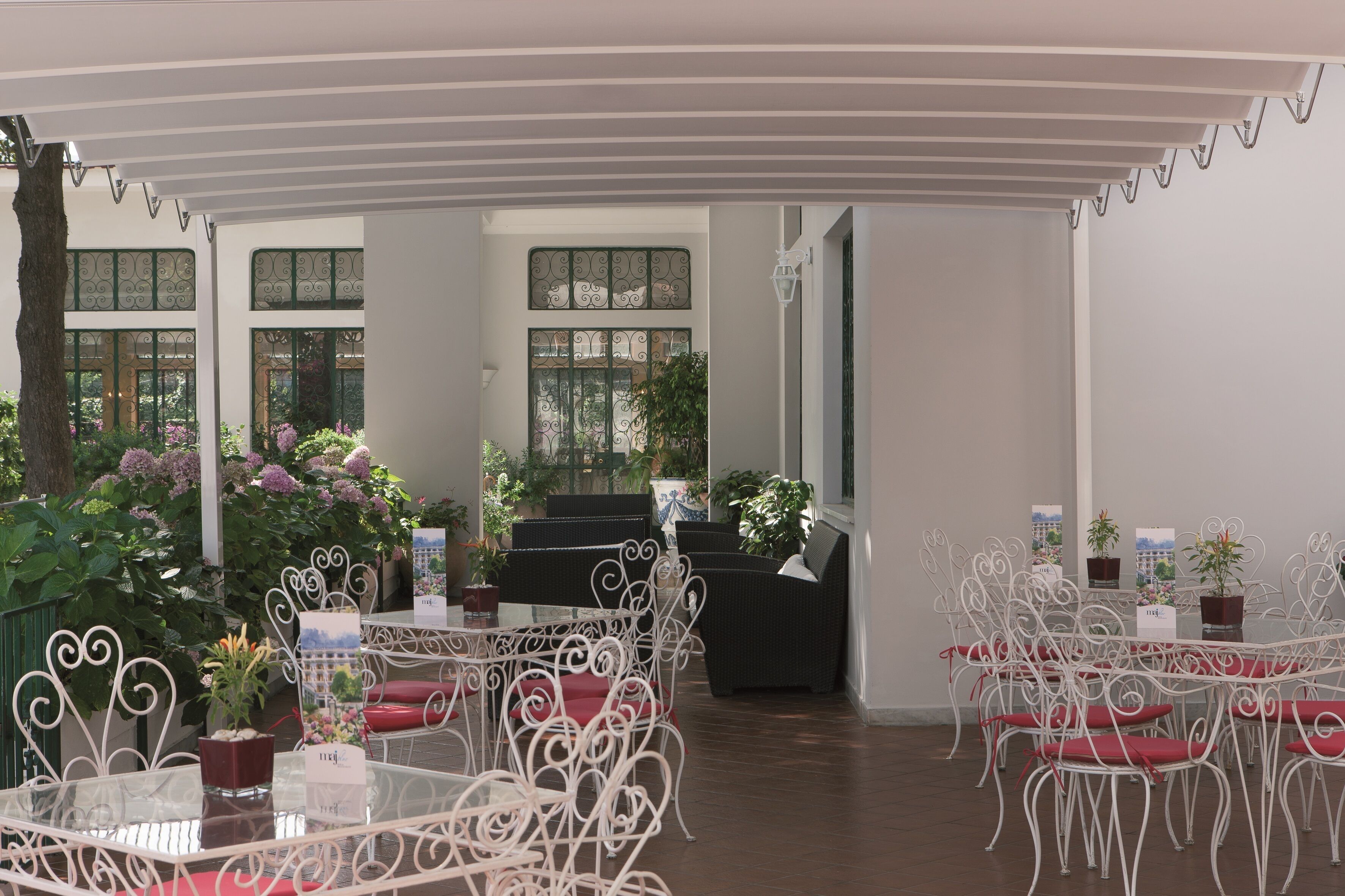 An indoor dining area with white metal chairs and tables, surrounded by potted plants and a piano.