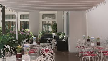 An indoor dining area with white metal chairs and tables, surrounded by potted plants and a piano.