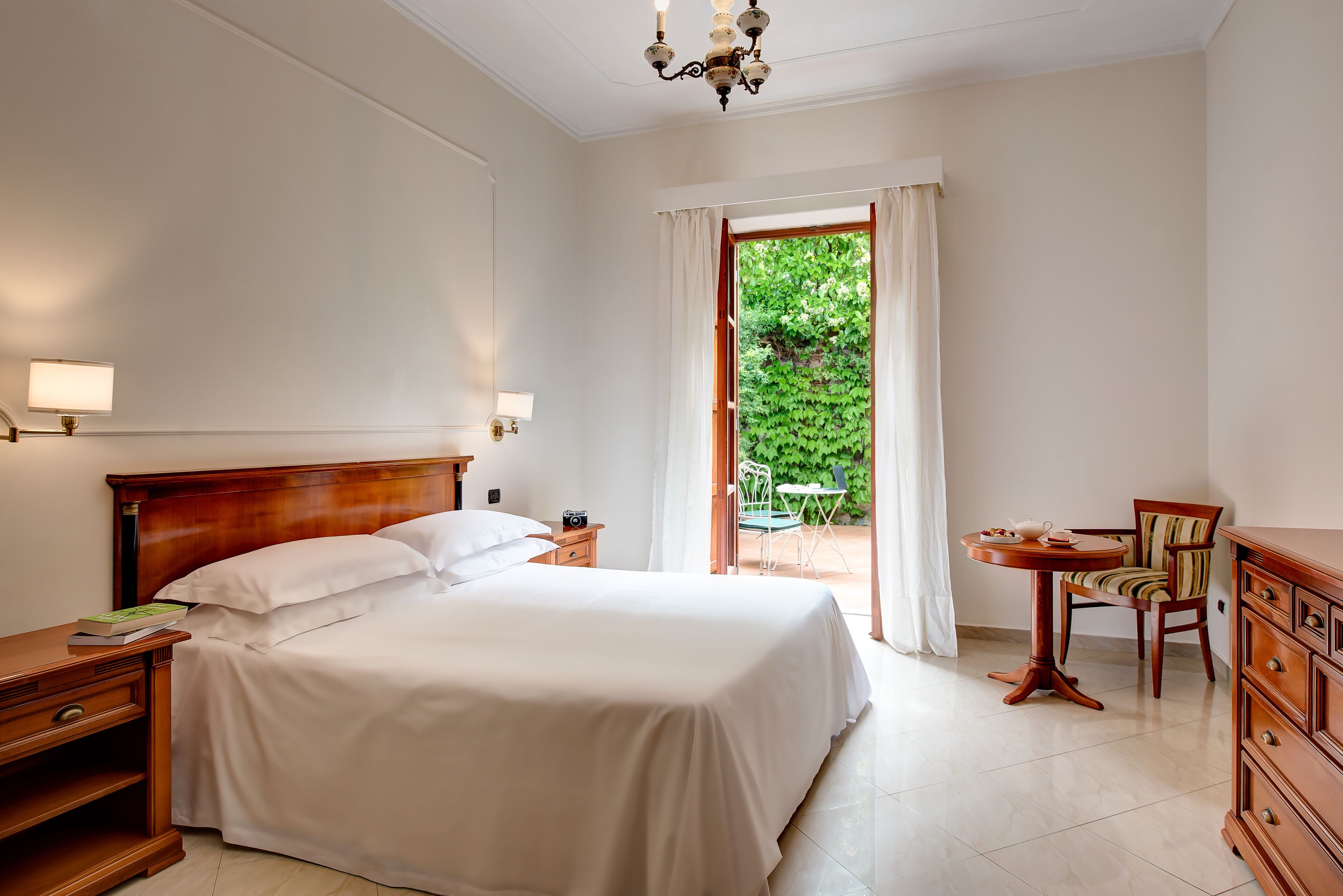 A bedroom with a bed, bedside table, lamp, and a view of a garden through a window.