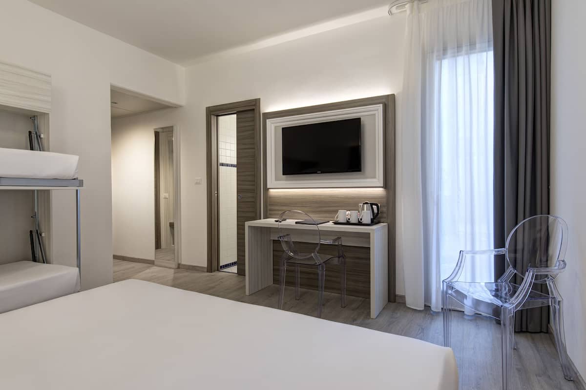 A modern hotel room with a bed, a desk with a TV, and two clear chairs.