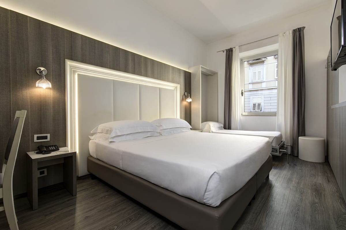 A modern hotel room with a large bed, bedside tables, a desk, and a window with curtains.
