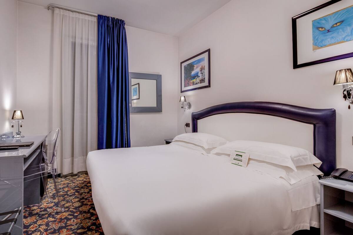 A hotel room with a bed, a desk with a chair, a TV, a window with curtains, and a framed picture on the wall.