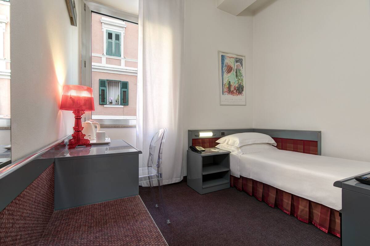 A hotel room with a single bed, a bedside table with a lamp, a small nightstand, a window with curtains, and a painting on the wall.
