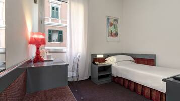 Ein Hotelzimmer mit einem Einzelbett, einem Nachttisch mit Lampe, einem kleinen Beistelltisch, einem Fenster mit Vorhängen und einem Bild an der Wand.