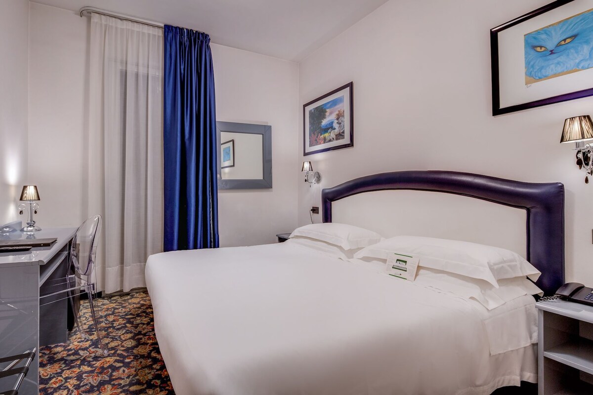 A hotel room with a bed, a desk with a chair, a TV, a window with curtains, and a framed picture on the wall.