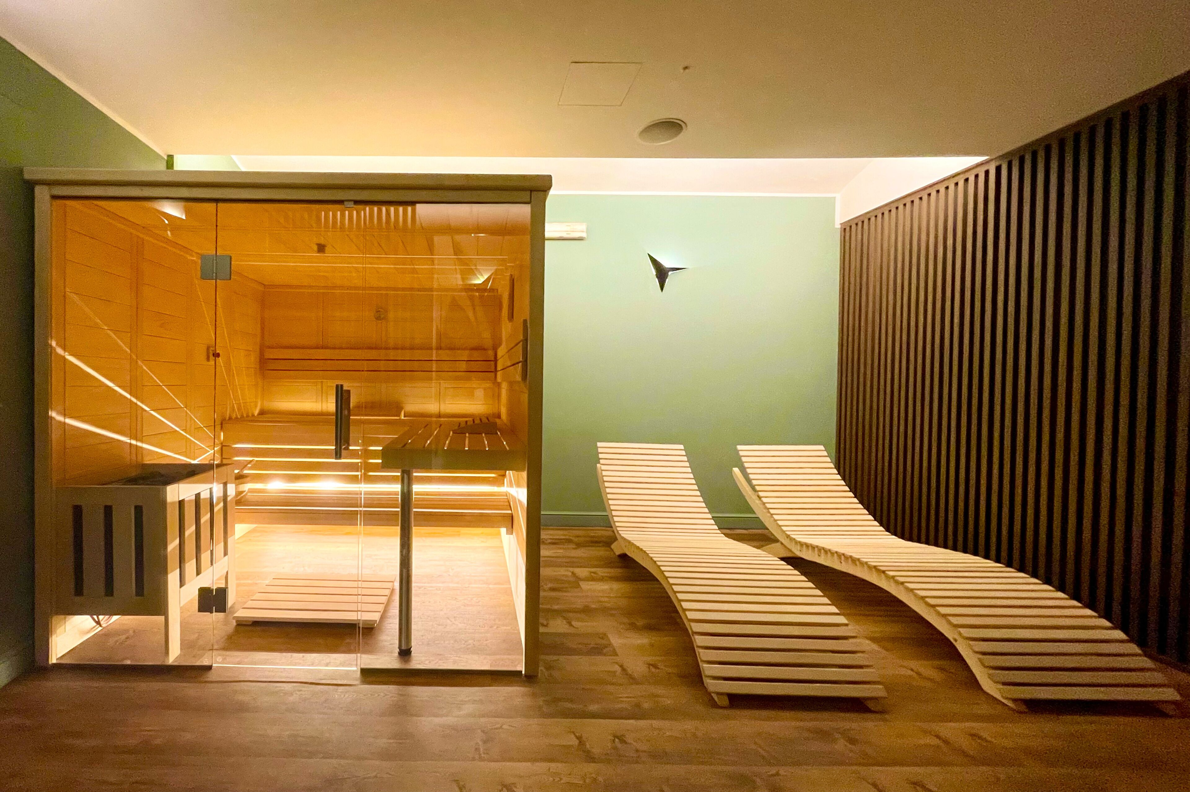 A sauna room with wooden benches and a glass door, a wooden bench outside the sauna, and a wall-mounted light fixture.