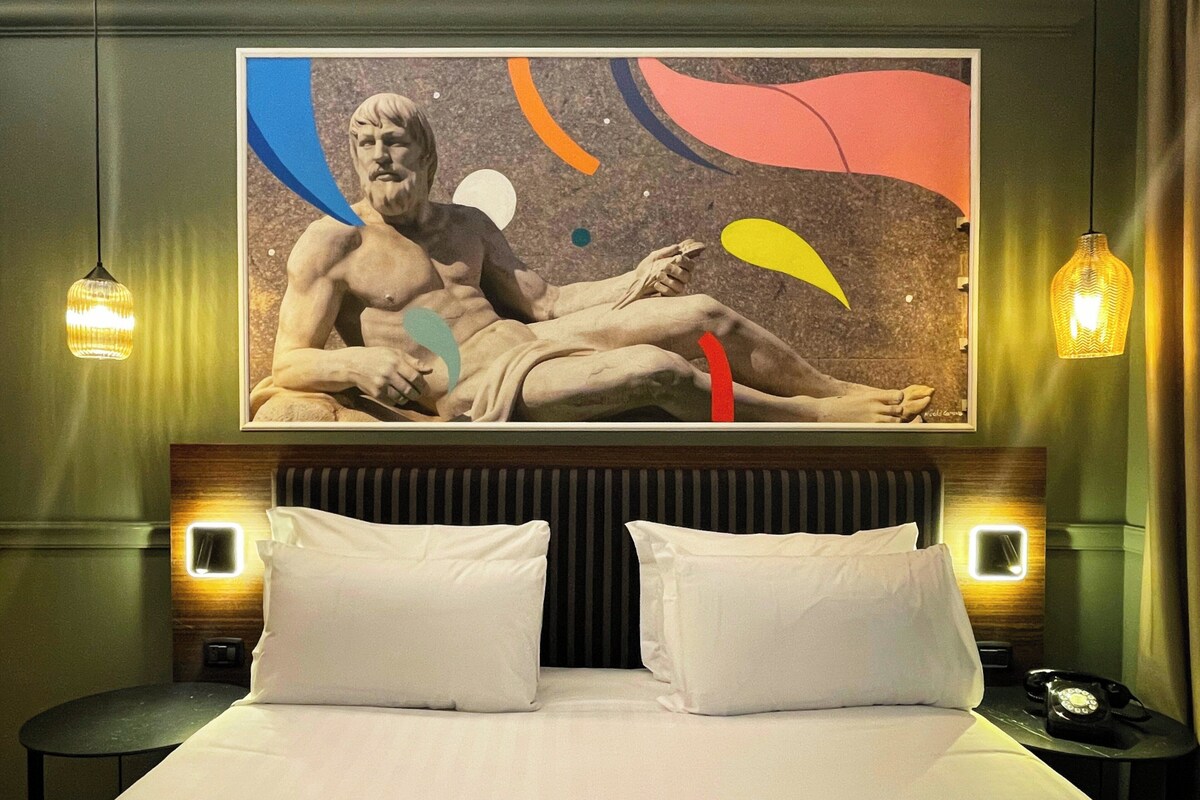 Habitación de hotel con una cama grande, dos almohadas, una mesita de noche con un teléfono y un cuadro colgado en la pared de una figura reclinada.
