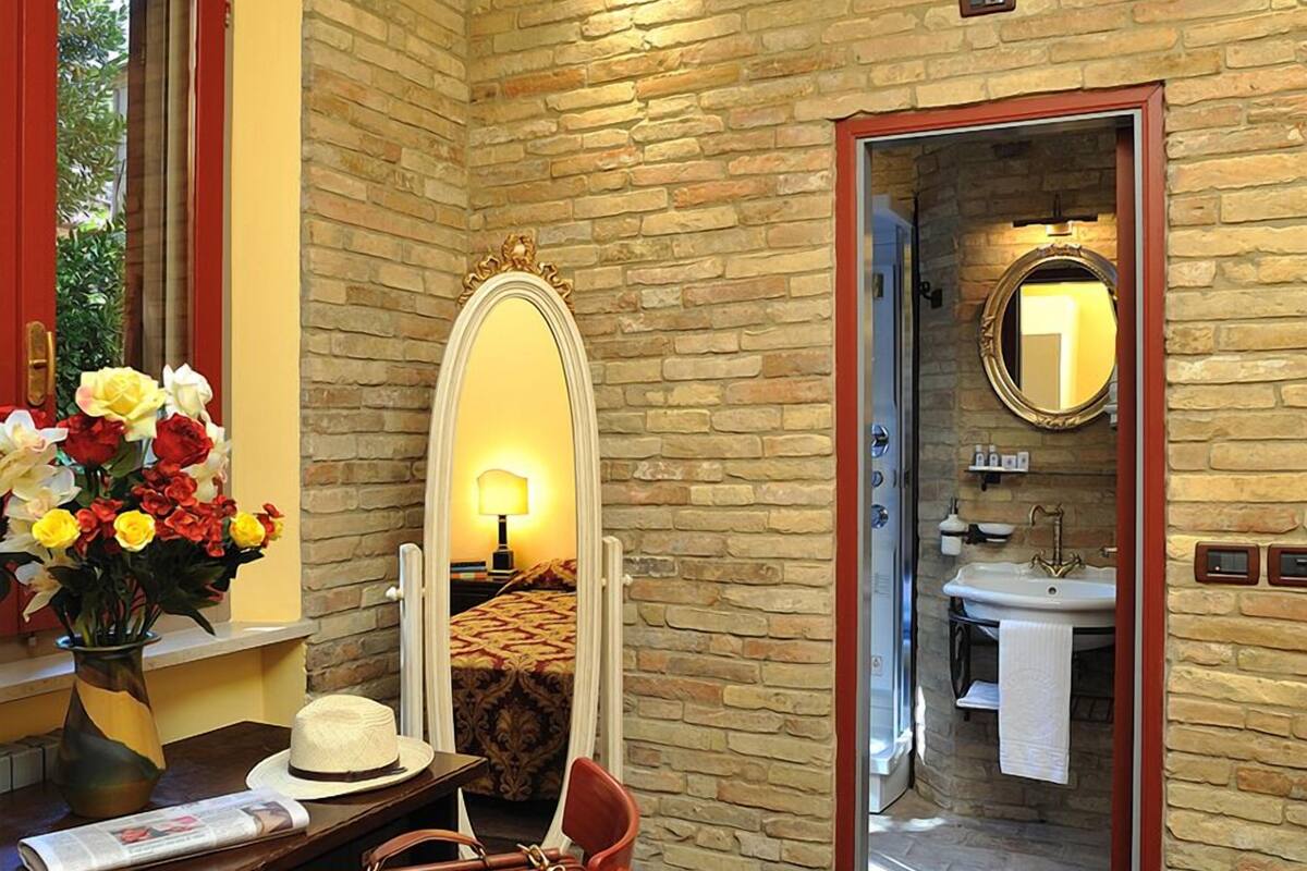 A room with a brick wall, a mirror, a bed, a desk with a chair, and a bathroom visible through an open door.