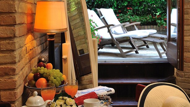 Una mesa de desayuno con fruteros, tetera, tazas y pasteles en una bandeja, con vistas a un jardín y una zona de estar al aire libre.