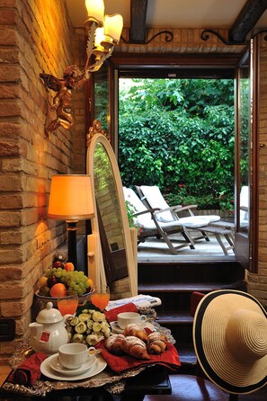 Una mesa de desayuno con fruteros, tetera, tazas y pasteles en una bandeja, con vistas a un jardín y una zona de estar al aire libre.
