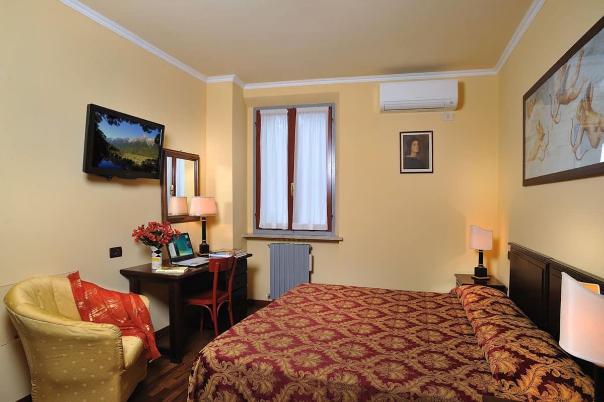 A hotel room with a bed, a desk with a computer, a chair, a window with curtains, and artwork on the walls.