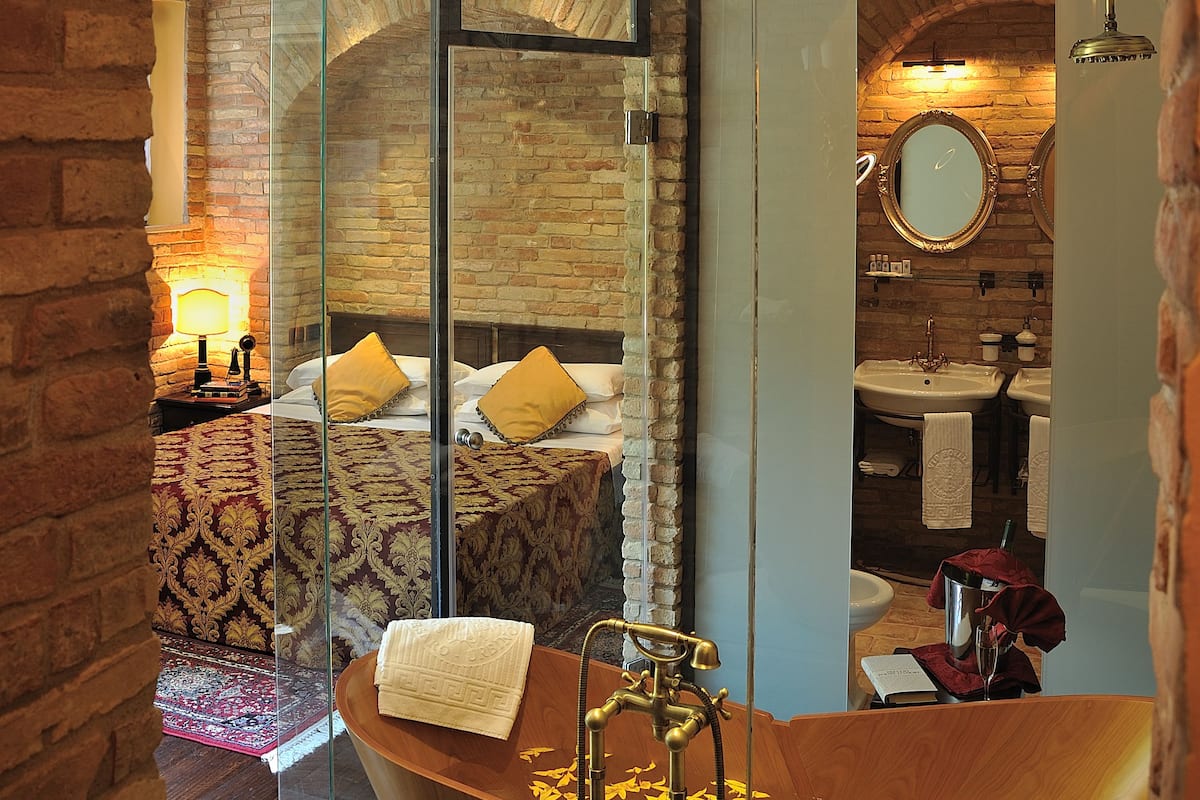 A modern bathroom with a large bathtub, a glass-enclosed shower, and a visible bed with a patterned rug.