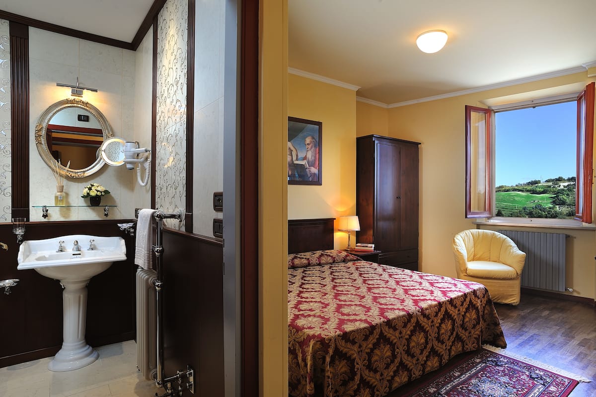 A hotel room with a bathroom, a bed with a patterned bedspread, a chair, and a window with a view.