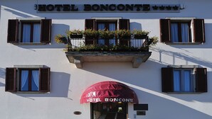 Un hotel llamado "HOTEL BONCONTE" con toldo rojo, varias ventanas con contraventanas y plantas en macetas en el balcón.