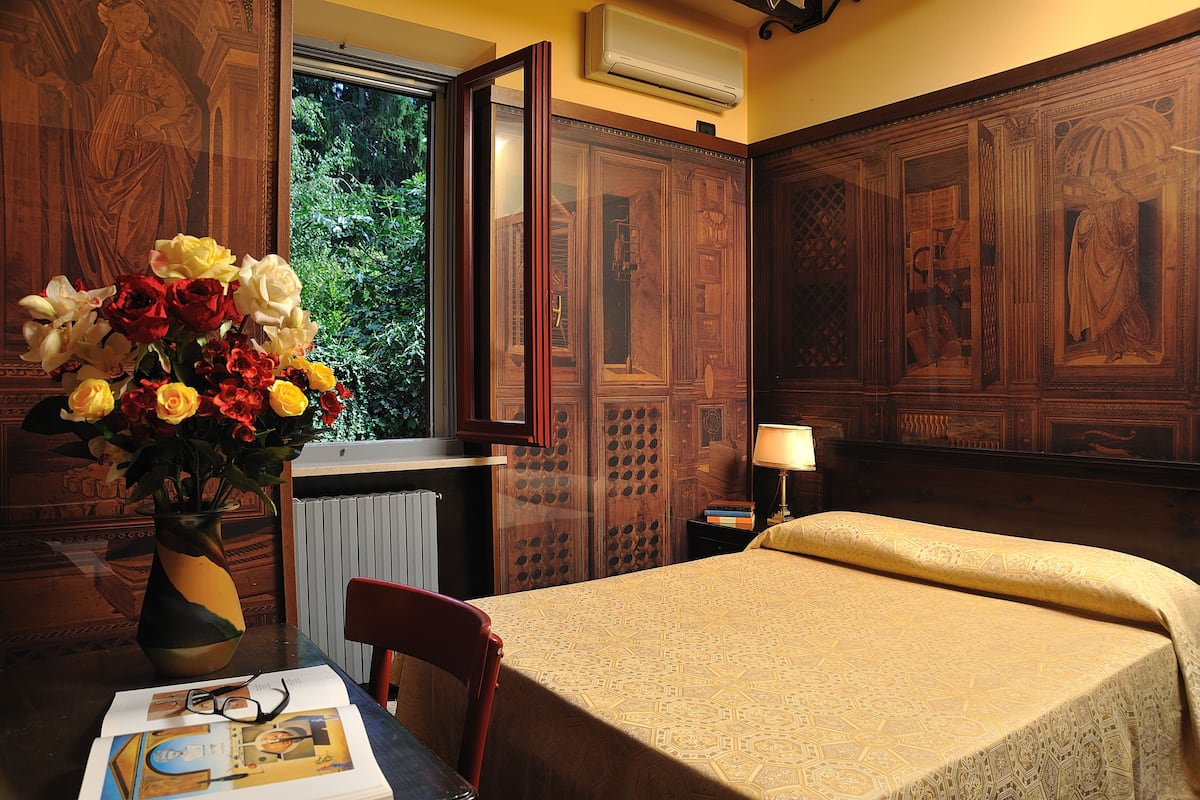 A room with a bed, a wooden cabinet, a vase with flowers, a table with a book and glasses, and a window with a view of greenery.