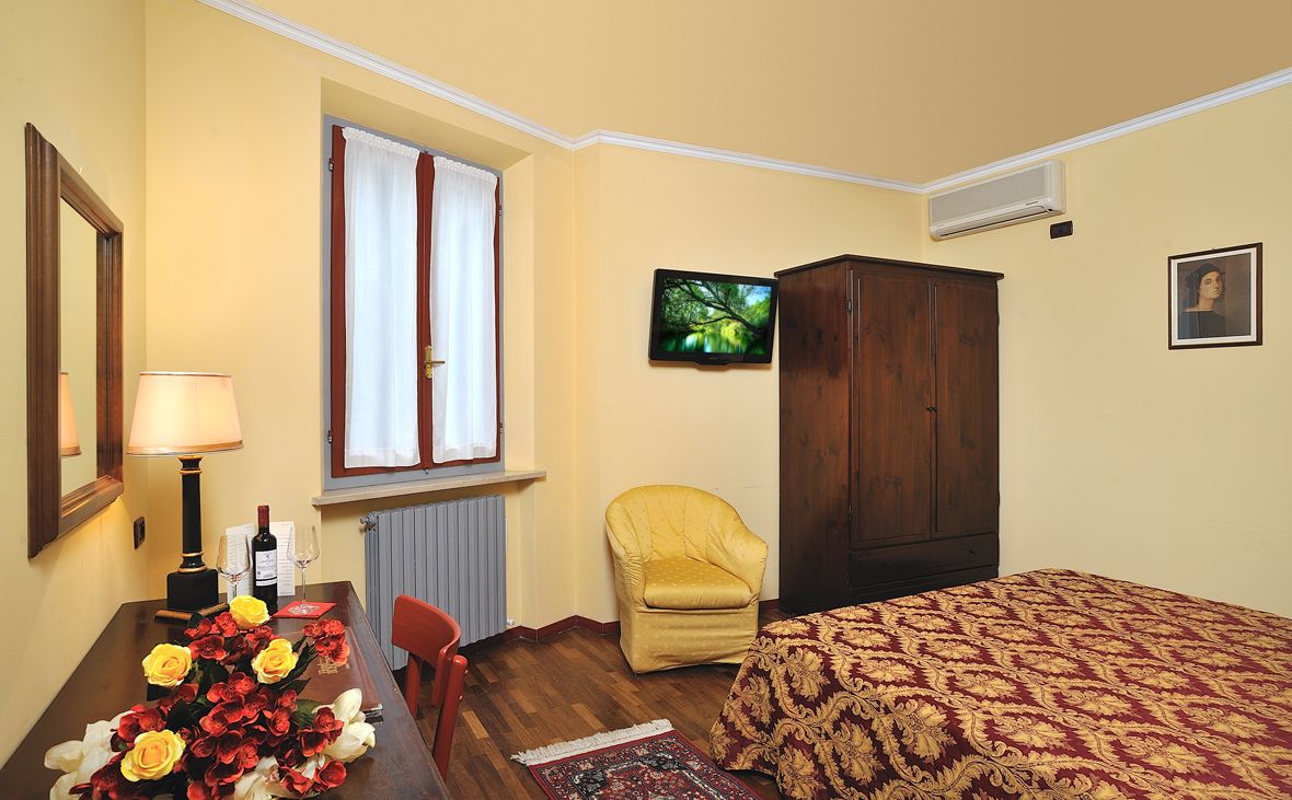 A room with a bed, a wooden wardrobe, a chair, a table with a lamp, and a window with curtains.