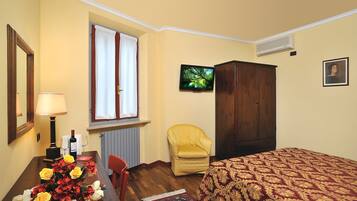 A room with a bed, a wooden wardrobe, a chair, a table with a lamp, and a window with curtains.