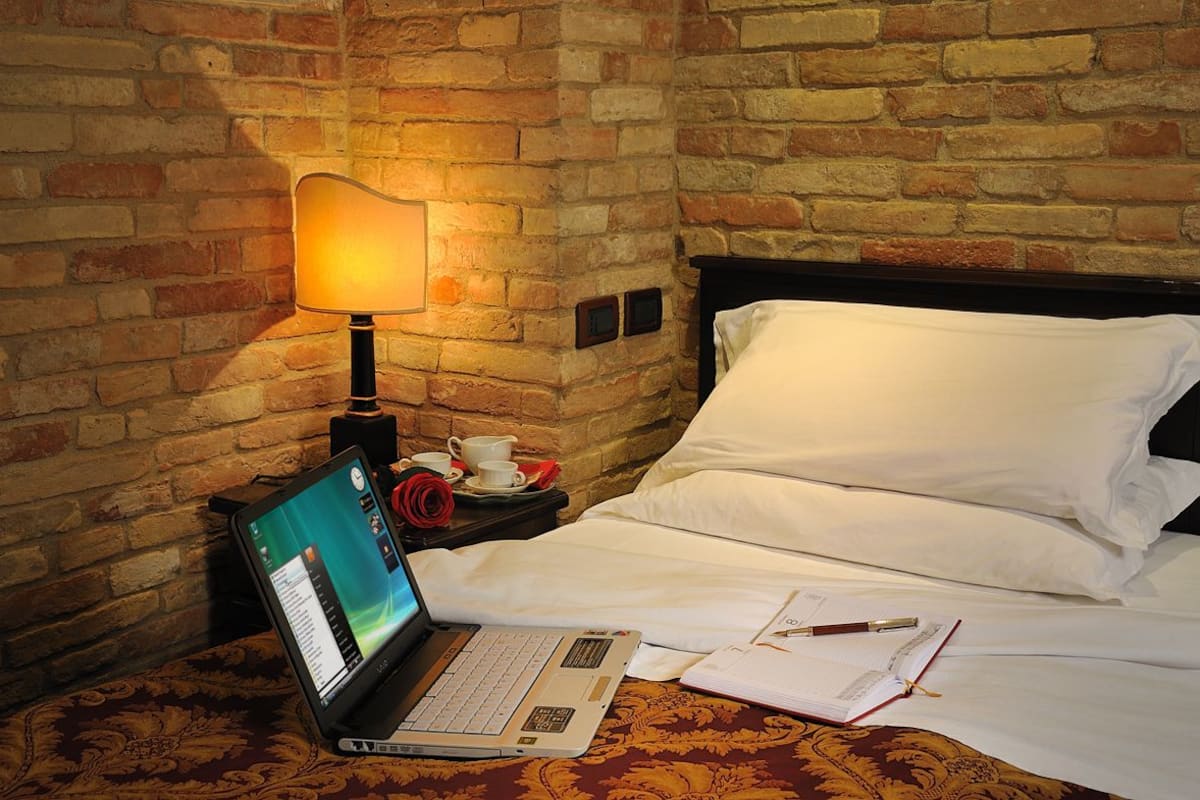 A hotel room with a brick wall, a bed with white linens, a laptop on the bedside table, a lamp, and a cup of coffee.