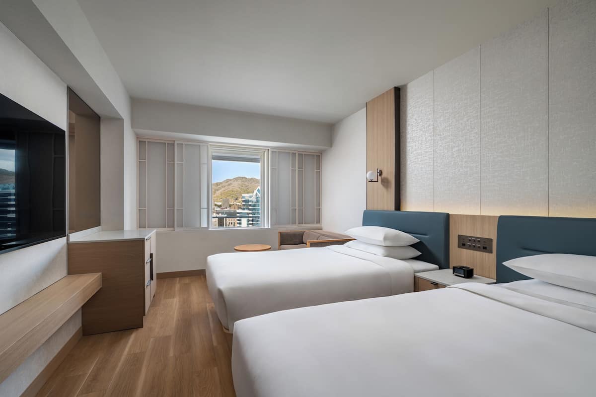 Habitación de hotel con dos camas, una mesita, una ventana y televisor integrado, todo con vistas a la ciudad.