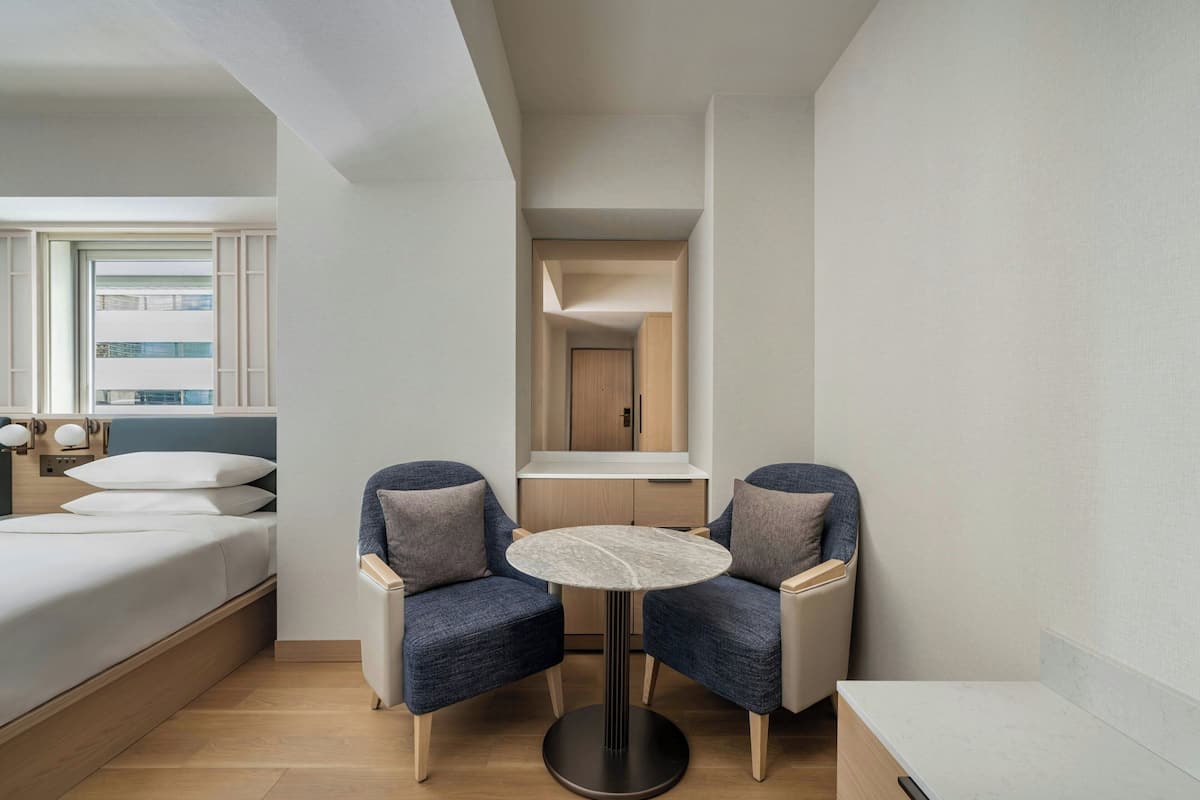 Habitación de hotel moderna con cama, dos sillones, una mesita redonda y vistas a la ciudad a través de una ventana.