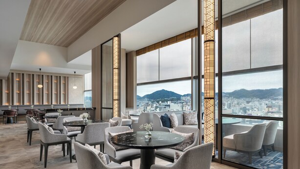 Un hall de hotel espacioso con techos altos, amplias ventanas que ofrecen vistas a la ciudad y mobiliario moderno.