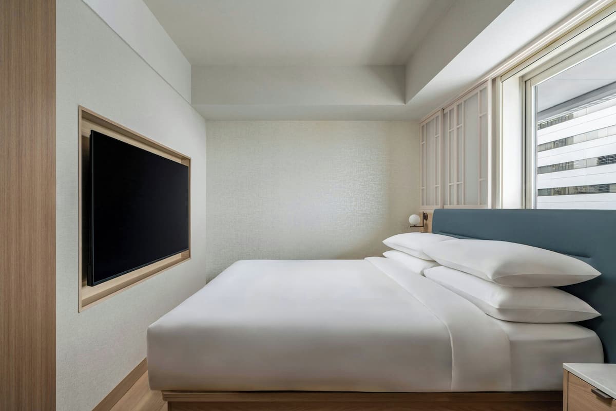 Una cama bien hecha con sábanas blancas, un televisor de pantalla plana montado en la pared y una ventana con persianas.