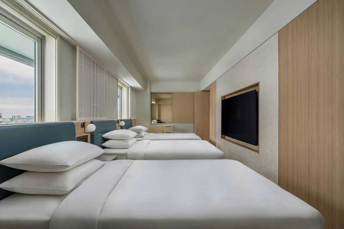 Una habitación de hotel moderna con tres camas, televisión, escritorio y ventana con vista a la ciudad.
