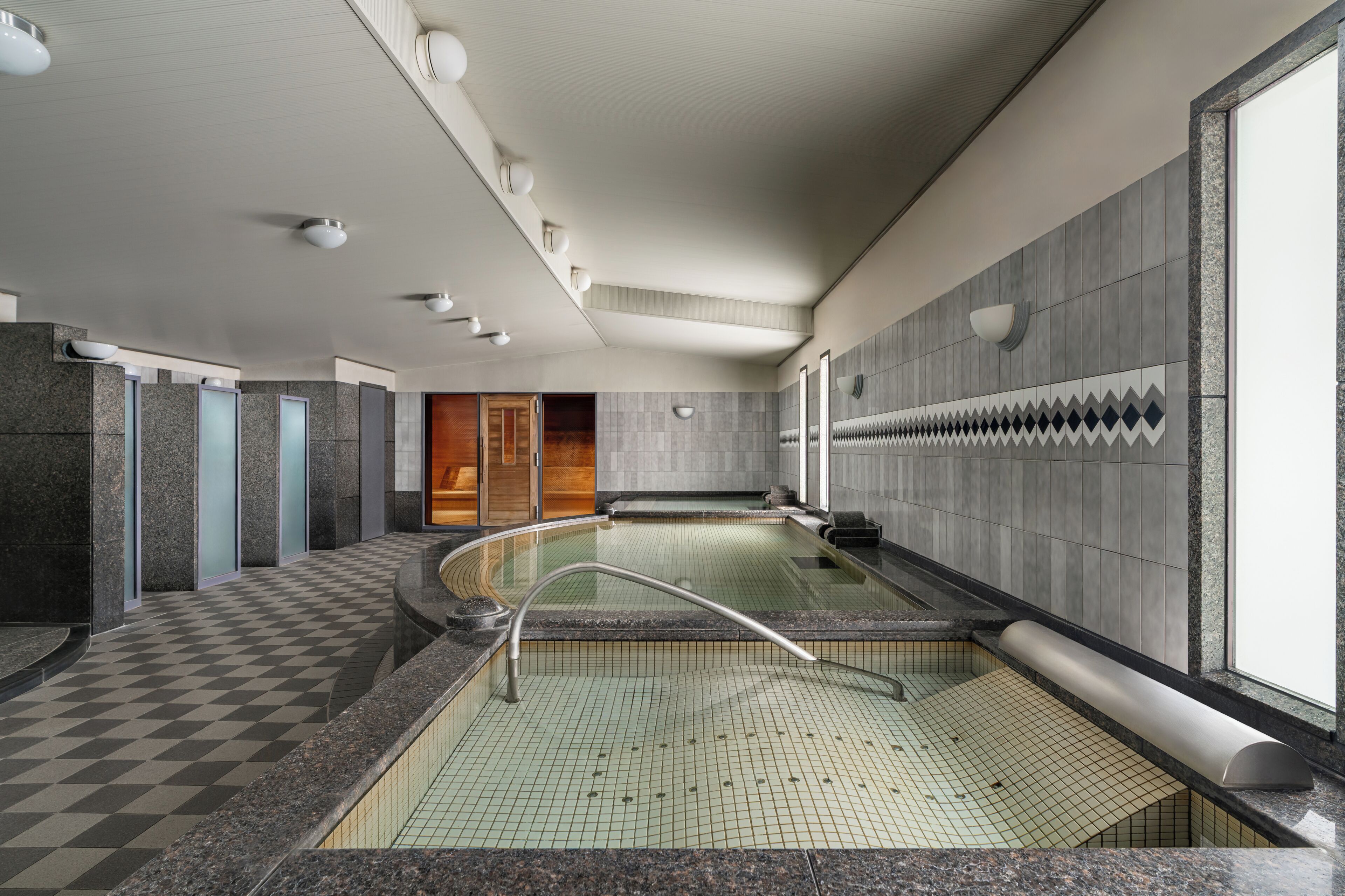 A modern indoor swimming pool with a checkered floor, a glass barrier, and a wooden door in the background.