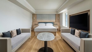 Una habitación de hotel moderna con una cama grande, dos sofás, una mesita redonda y un televisor grande.