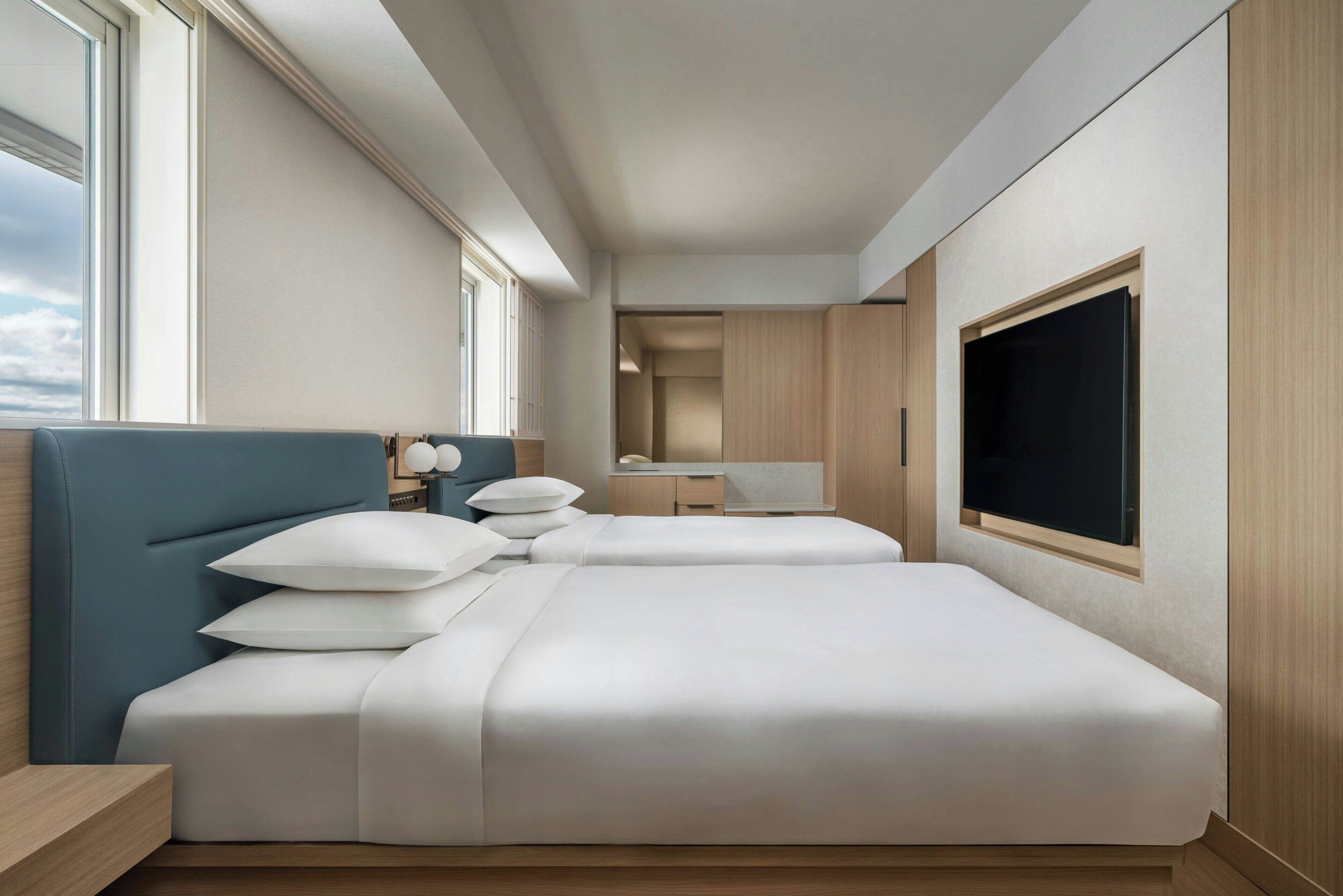 A modern hotel room with two beds, a large window, a TV, and a wooden cabinet.