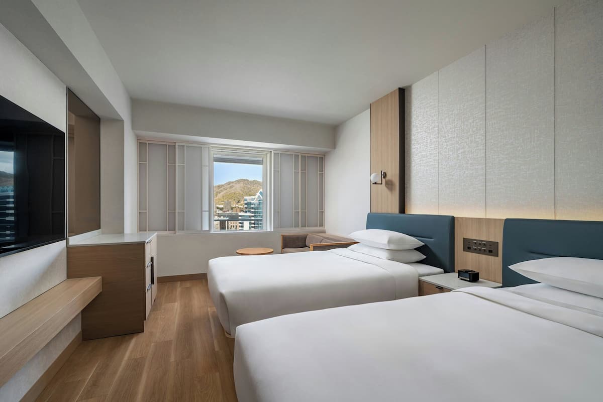 Una habitación de hotel moderna con dos camas, un escritorio, una mesita redonda y una ventana con vista a la ciudad.