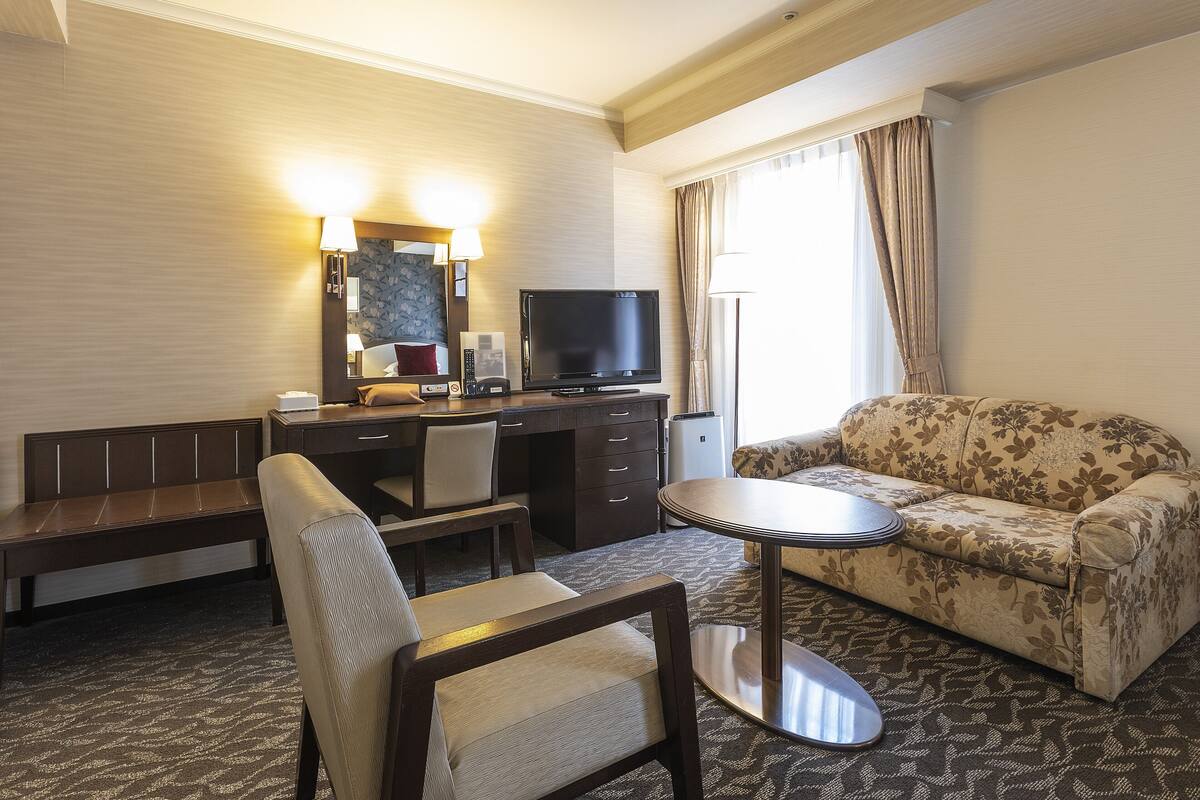 A hotel room with a bed, a desk with a TV, a chair, a sofa, and a small table.