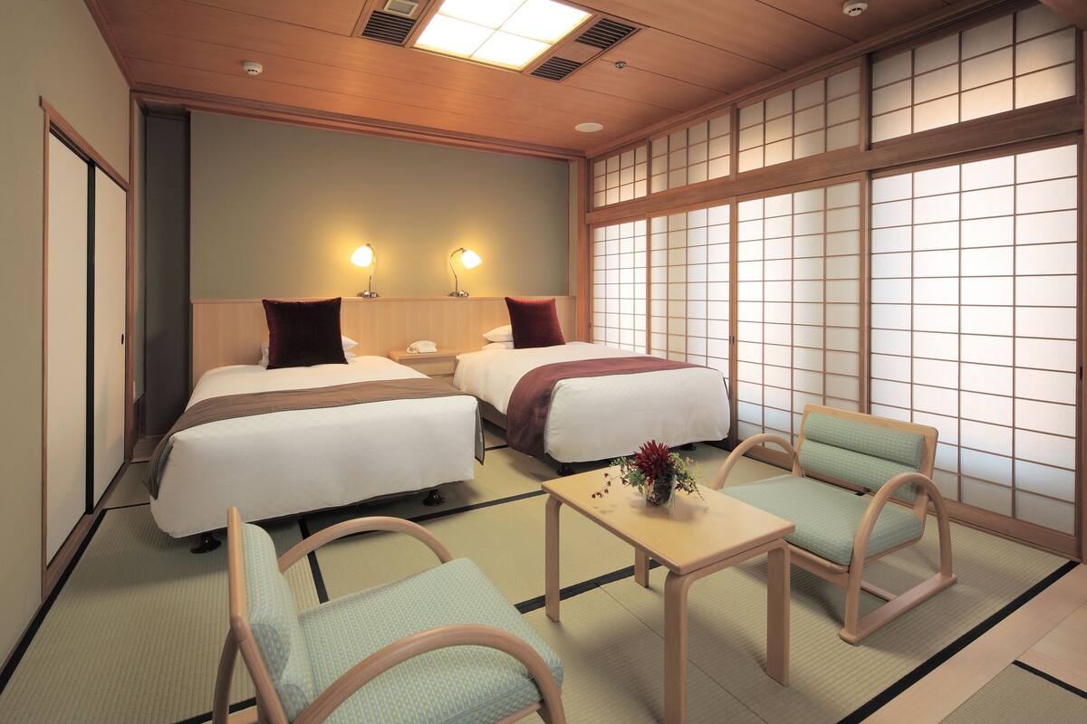 A hotel room with two beds, a small table with a vase, and traditional Japanese-style chairs.