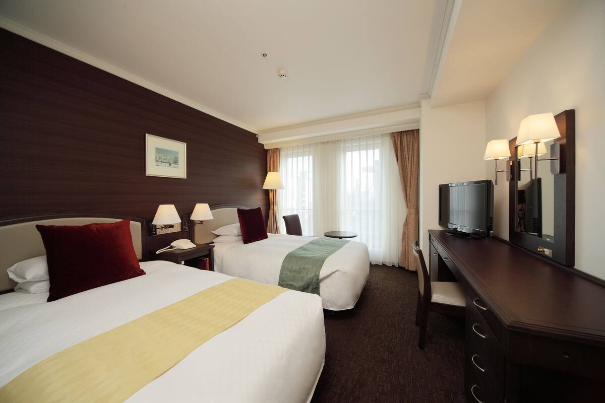 A hotel room with two beds, a desk, a television, and a window with curtains.