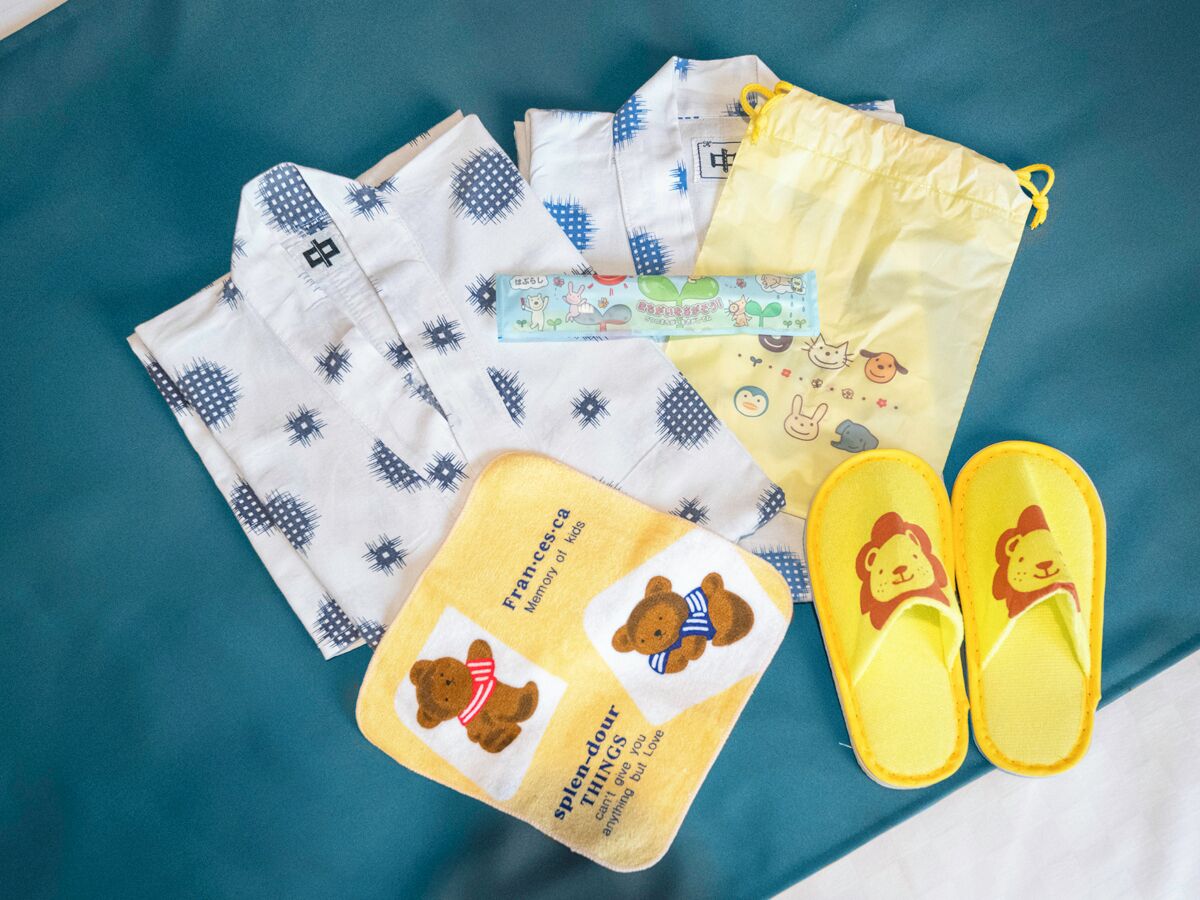 Un lit avec un drap bleu, un sac jaune à cordon, une paire de pantoufles jaunes et un oreiller blanc orné d’un motif bleu.