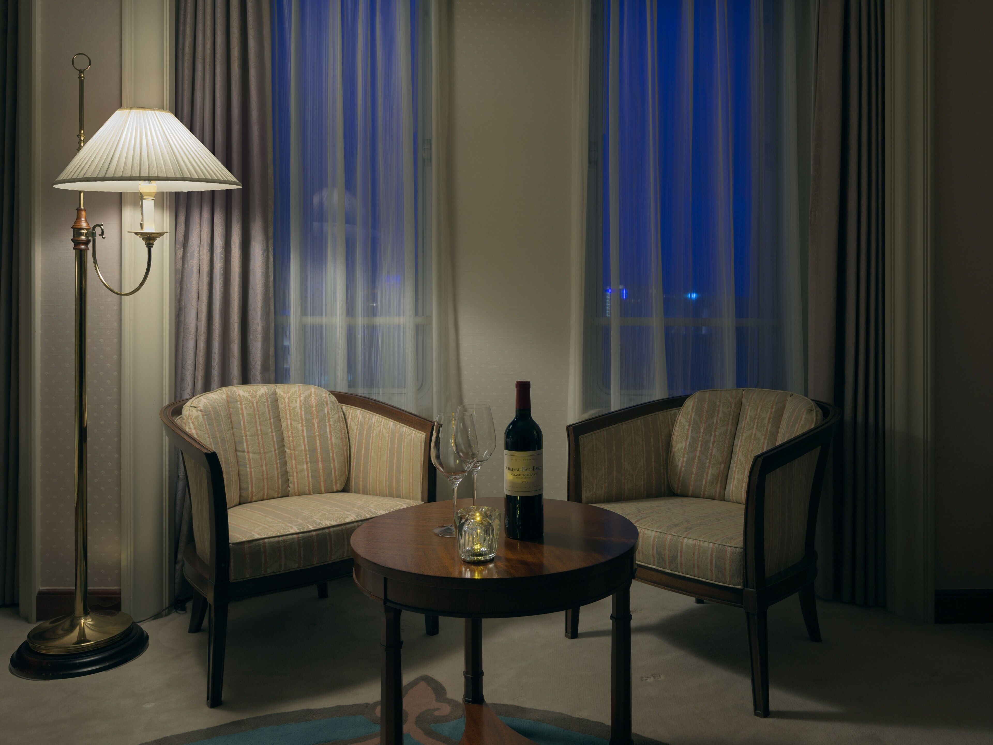 Habitación de hotel con dos sillones, una mesa redonda, una lámpara de pie y una botella de vino.
