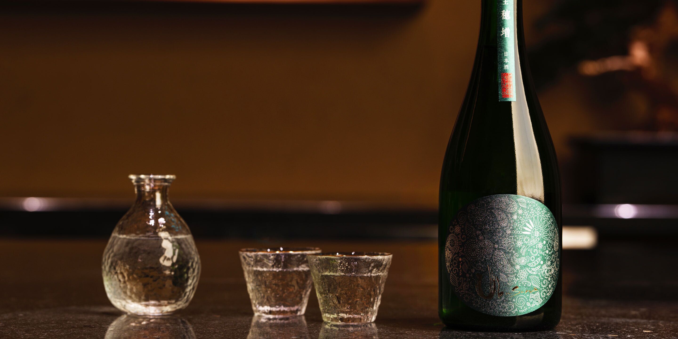 A bottle of sake with a decorative label, a clear glass vase, and two small glasses on a reflective surface.