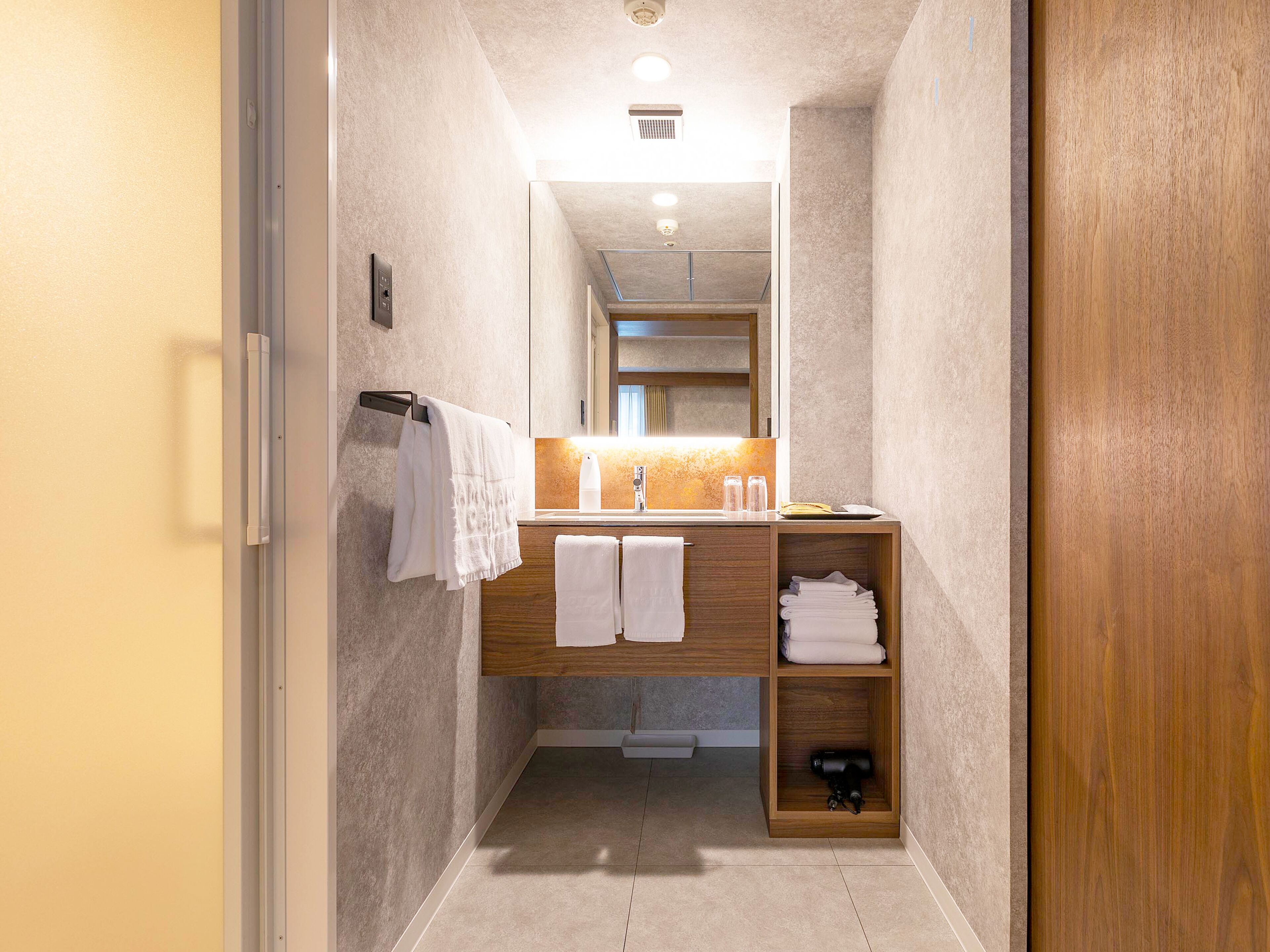 A narrow hallway leading to a bathroom with a wooden vanity, two towels hanging on the wall, and a shelf with folded towels.
