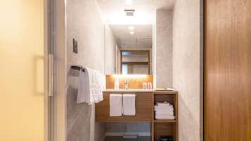 A narrow hallway leading to a bathroom with a wooden vanity, two towels hanging on the wall, and a shelf with folded towels.