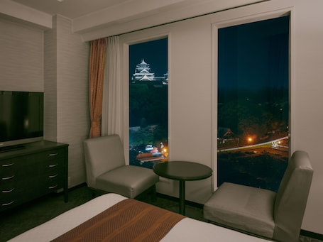 A hotel room with a bed, two armchairs, a small table, a TV, and a large window with a view of a castle and city lights.. Kumamoto Hotel Castle