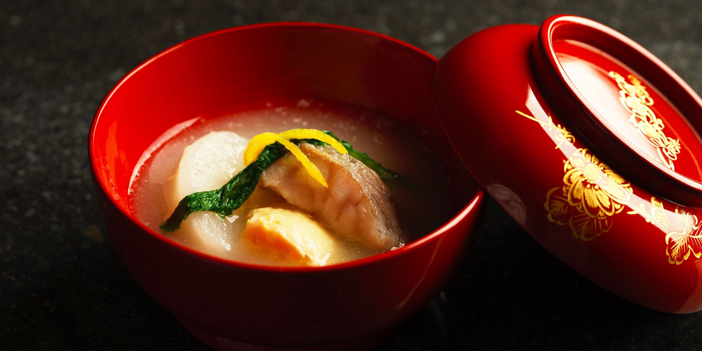A red bowl with a soup containing vegetables and meat, placed on a dark surface.
