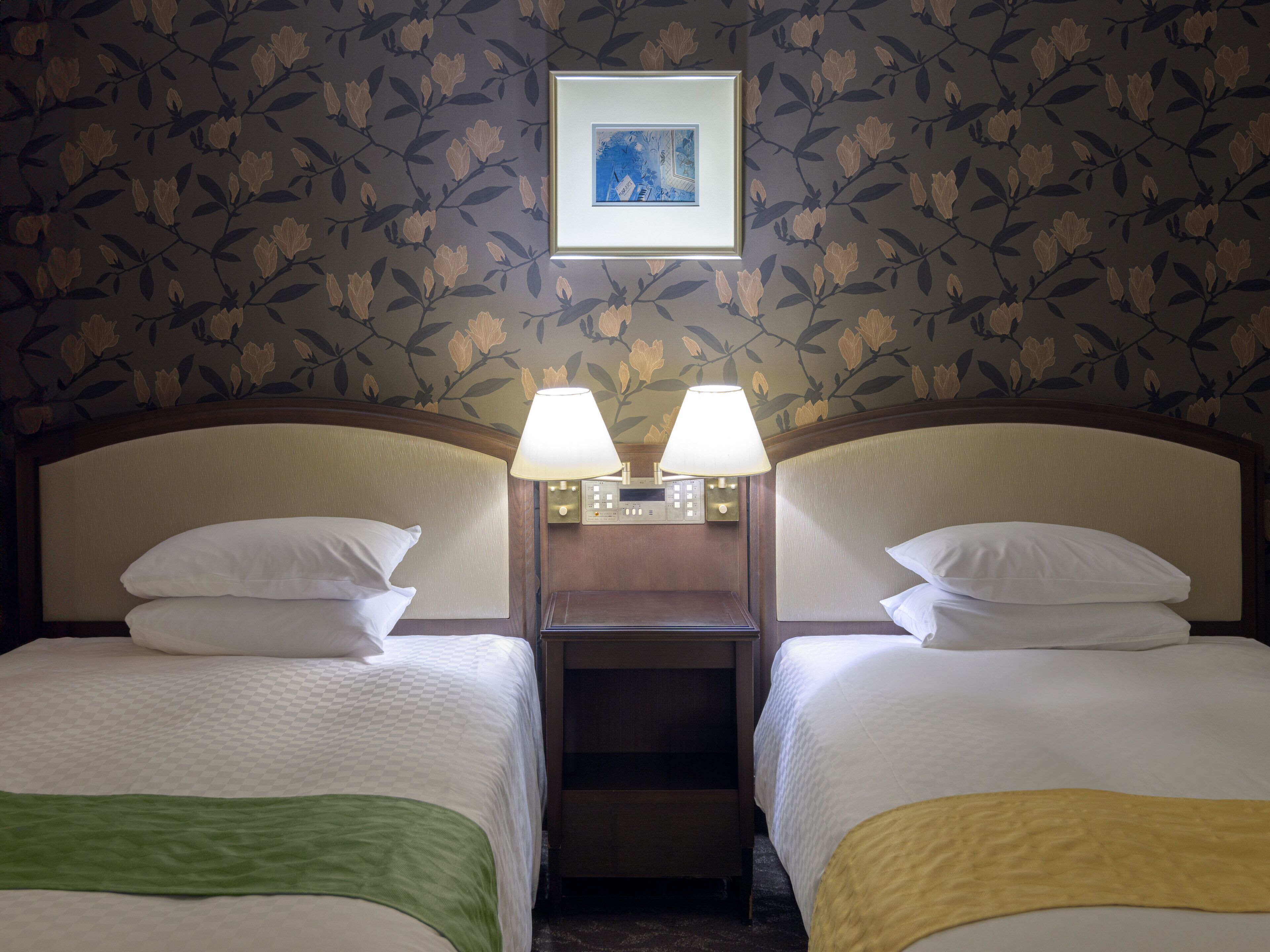 A hotel room with two beds, a nightstand with lamps, a framed picture on the wall, and floral wallpaper.