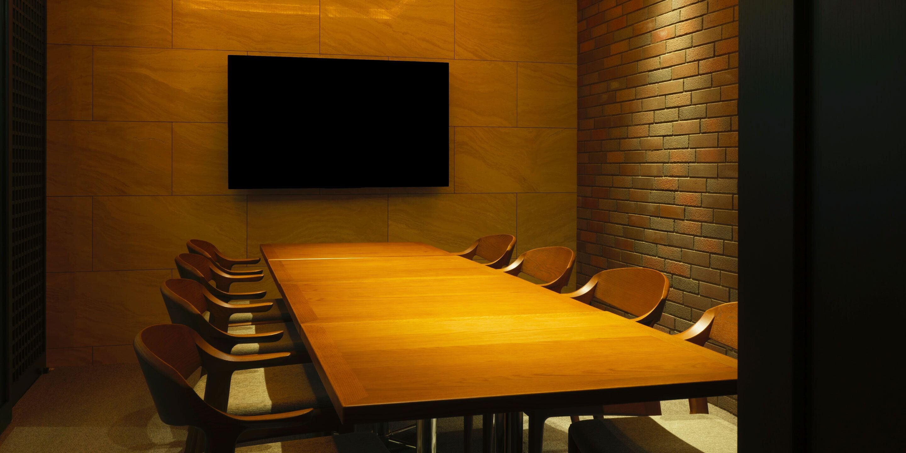 A conference room with a long wooden table and multiple chairs, a flat-screen TV mounted on the wall, and a brick wall on one side.