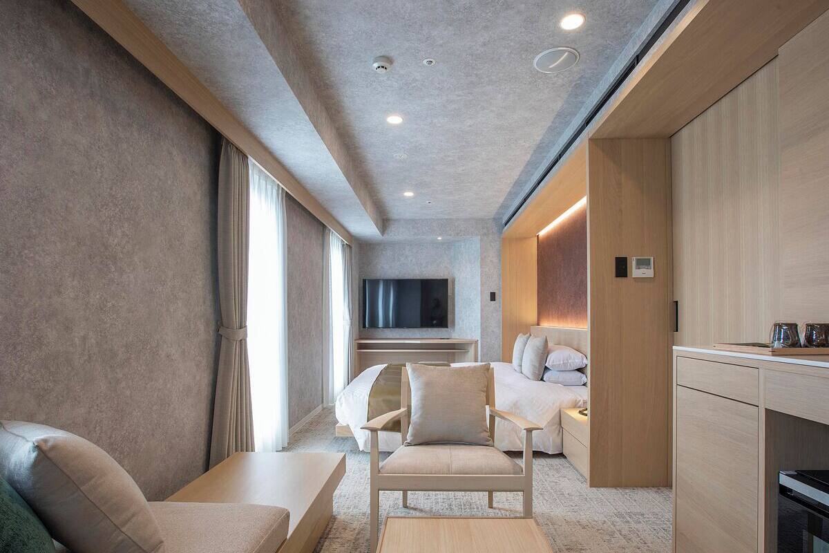 A modern hotel room with a sofa, armchair, and a flat-screen TV mounted on the wall.