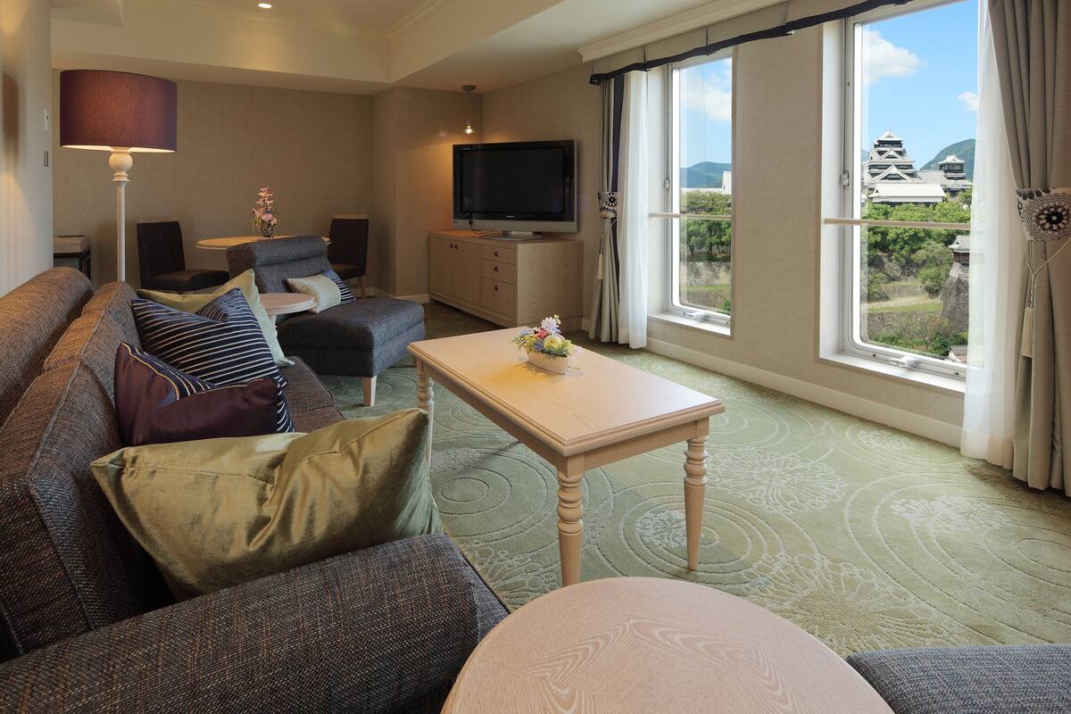 A modern living room with a sofa, armchair, and a small table with a vase. A television is mounted on the wall, and there is a window with a view of traditional buildings.