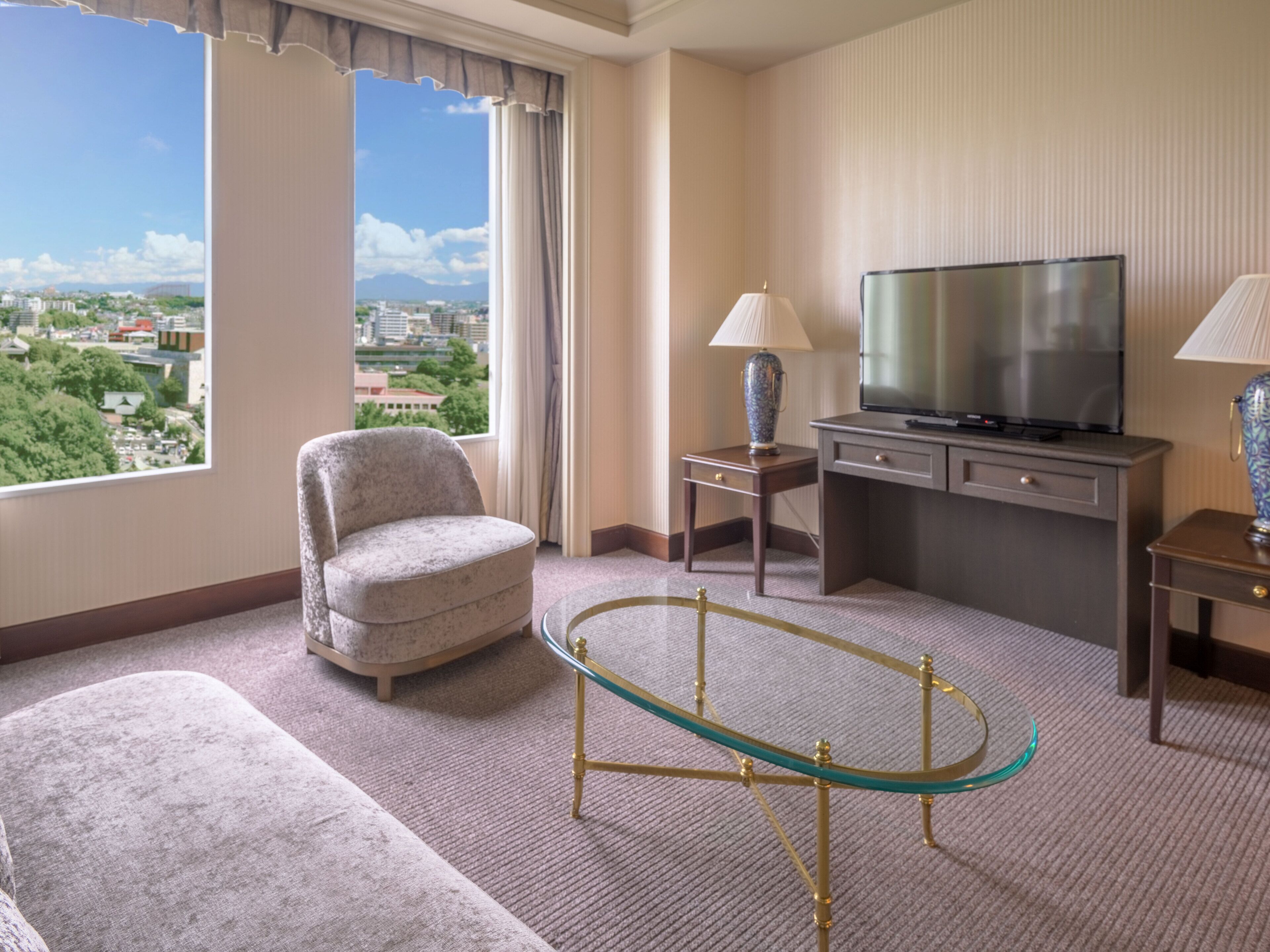 A hotel room with a large window, a television, a sofa, a glass coffee table, and two lamps.